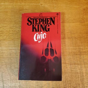 May include: A mass market paperback book titled Cujo by Stephen King. The cover features a vibrant red background that fades into black at the bottom. The text reads From the author of IT, Stephen King, and Cujo. A dark paw print is visible.
