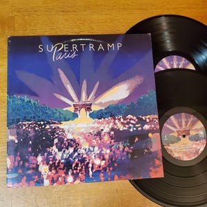 May include: The album cover for Supertramp's "Paris" album features a vibrant illustration of a crowd scene with the Arc de Triomphe. Two black vinyl records are visible, one partially obscuring the other. The album cover is square and has the band's name and album title.