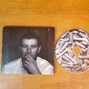 May include: A CD and its case are displayed on a wooden surface. The CD case features a black and white photo of a person smoking a cigarette. The CD itself has a design of cigarette butts.