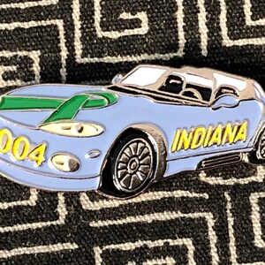 May include: A blue and silver car pin with a green ribbon on the hood. The car has "2004" and "INDIANA" written on it.