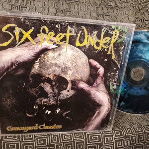 May include: A CD case with a black and white image of a hand holding a skull. The text "Six Feet Under" is in yellow and the text "Graveyard Classics" is in white.