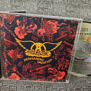 May include: A CD case with a black background and red, white, and black graphics. The case features the band Aerosmith and the album title "Permanent Vacation".