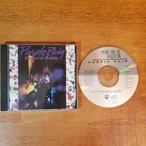 May include: A CD and its case are displayed on a wooden surface. The CD case features the album title "Purple Rain" and the artist's name. The CD itself has the text "Prince and the Revolution" and "Purple Rain."
