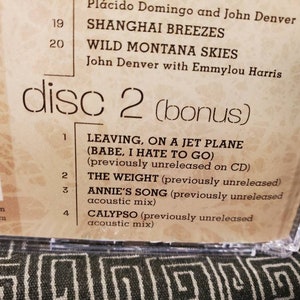 May include: A close-up of a CD case back cover featuring a tracklist for disc 2. Visible text includes Shanghai Breezes, Wild Montana Skies, Leaving on a Jet Plane, The Weight, Annie's Song, and Calypso, with notes about previously unreleased tracks.