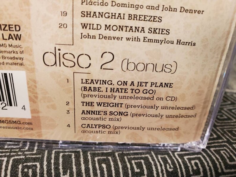 May include: A close-up of a CD case back cover featuring a tracklist for disc 2. Visible text includes Shanghai Breezes, Wild Montana Skies, Leaving on a Jet Plane, The Weight, Annie's Song, and Calypso, with notes about previously unreleased tracks.