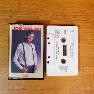 K&ouml;nnte beinhalten: Zwei Vintage-Musikkassetten auf einer Holzoberfl&auml;che. Die erste Kassettenh&uuml;lle zeigt ein Foto von Joe Walsh und den Text "THE BEST OF JOE WALSH". Die zweite Kassette ist wei&szlig; mit schwarzem Text, darunter "ROCKY MOUNTAIN WAY" und "MCA Records Inc."
