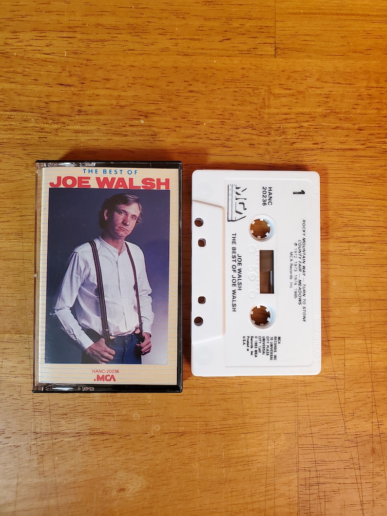 K&ouml;nnte beinhalten: Zwei Vintage-Musikkassetten auf einer Holzoberfl&auml;che. Die erste Kassettenh&uuml;lle zeigt ein Foto von Joe Walsh und den Text "THE BEST OF JOE WALSH". Die zweite Kassette ist wei&szlig; mit schwarzem Text, darunter "ROCKY MOUNTAIN WAY" und "MCA Records Inc."