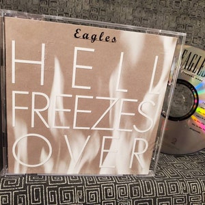 May include: A compact disc case with a white background and the text "Eagles Hell Freezes Over" on the spine. The front of the case features a white text overlay on a brown background with the text "Eagles" and "Hell Freezes Over". The CD is visible in the case and has a rainbow iridescent effect.