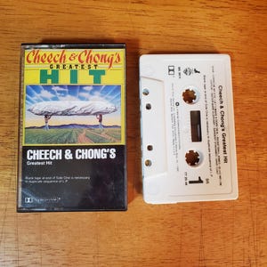 May include: A Cheech & Chong's Greatest Hit cassette tape with its plastic case. The cover features a large white cloud over a field with the title text. The white cassette shell displays the tracklist, the number 1, and Warner Bros. logos.