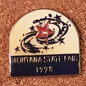 May include: A gold-colored pin with a white background and black and red design. The pin features a red star with a white stripe in a swirling black design. The text "MONTANA STATE FAIR 1998" is printed in black on a blue banner.