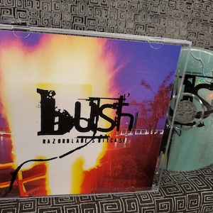 以下が含まれることがあります： 紫、オレンジ、黄色の背景のCDケース。バンド「Bush」が黒で印刷され、その下にアルバムタイトル「Razorblade Suitcase」が印刷されています。ケースの中にCDが見えます。