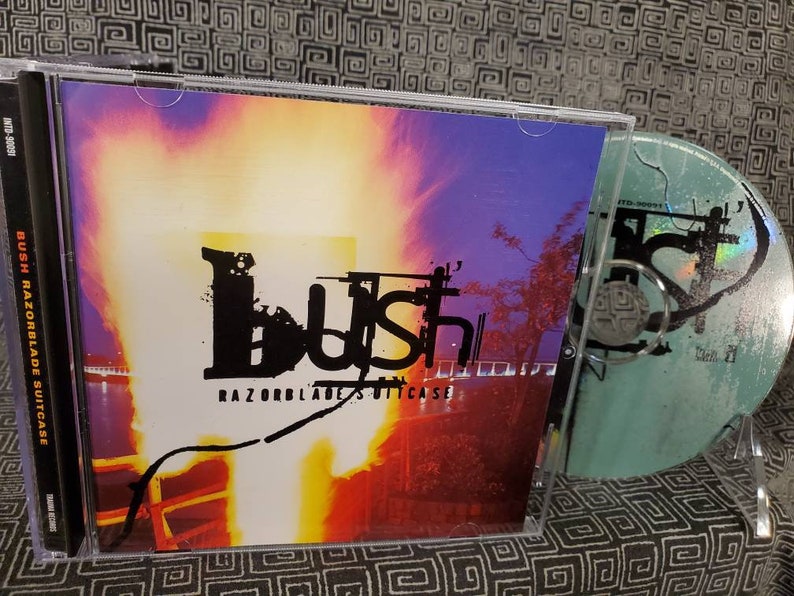 以下が含まれることがあります： 紫、オレンジ、黄色の背景のCDケース。バンド「Bush」が黒で印刷され、その下にアルバムタイトル「Razorblade Suitcase」が印刷されています。ケースの中にCDが見えます。