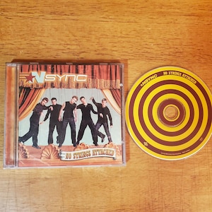 May include: An *NSYNC No Strings Attached CD in a clear plastic jewel case with a cover featuring the band members as marionettes on a stage. Beside the case is the circular disc, which has a yellow and brown bullseye pattern and text.