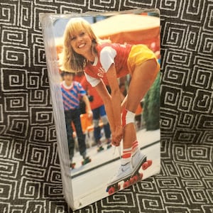 May include: A woman wearing a red and yellow outfit is roller skating. She is wearing white roller skates with red wheels. She is smiling and looking at the camera. The background is a blurry image of a street scene.