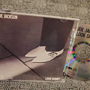 May include: A Joe Jackson "Look Sharp!" CD album. The album cover features a black and white image of a person's legs and white shoes. The CD is visible next to the case, with the album title and track list.