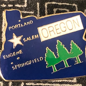May include: A blue and gold Oregon state pin. The pin features the state's outline, with the word "OREGON" in a white rectangle. It also includes the names of cities like Portland and Salem, a white star, and green trees.