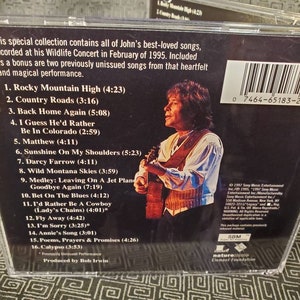 May include: A CD case with a photo of John Denver holding a guitar. The back of the case lists the songs on the CD, including "Rocky Mountain High", "Country Roads", and "Back Home Again".