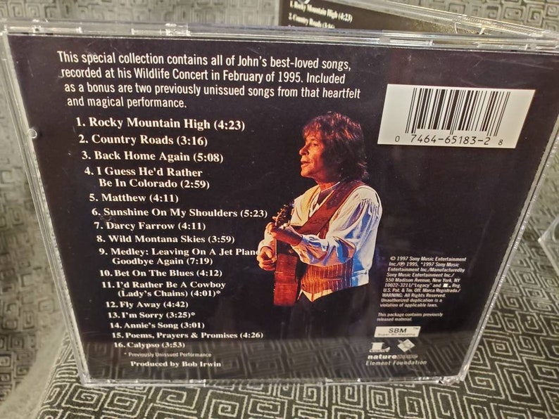 May include: A CD case with a photo of John Denver holding a guitar. The back of the case lists the songs on the CD, including "Rocky Mountain High", "Country Roads", and "Back Home Again".