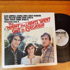 May include: Album cover for "The Night the Lights Went Out in Georgia" soundtrack. Features three people and the title in red and white text. A black vinyl record is next to the album cover. Performers include Glen Campbell, George Jones, and Tanya Tucker.