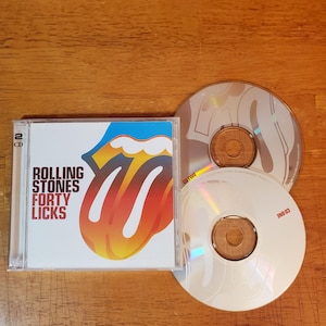 May include: A CD case with the Rolling Stones' "Forty Licks" album cover, featuring the band's iconic tongue and lips logo in red, orange, and blue. Two CDs are visible, one with the logo and the other with "CD ONE" printed on it.