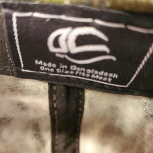 Peut inclure: &Eacute;tiquette noire et blanche avec le texte "Made in Bangladesh One Size Fits Most" et un logo stylis&eacute;. L'&eacute;tiquette est attach&eacute;e &agrave; une casquette de baseball camouflage noir et blanc.