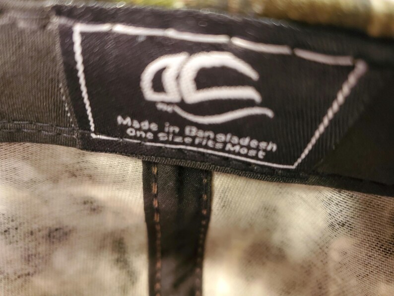 Peut inclure: &Eacute;tiquette noire et blanche avec le texte "Made in Bangladesh One Size Fits Most" et un logo stylis&eacute;. L'&eacute;tiquette est attach&eacute;e &agrave; une casquette de baseball camouflage noir et blanc.