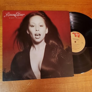 May include: Album cover for Yvonne Elliman's "Night Flight" album. The cover features a sepia-toned image of Elliman with long dark hair. The album cover is red with the album title and artist's name in white script. A black vinyl record is next to the album.