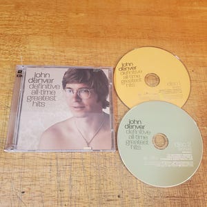 May include: A John Denver Definitive All-Time Greatest Hits 2-CD set in a clear plastic case. The cover features a portrait of John Denver wearing glasses and a necklace. Included are two discs, one yellow labelled disc 1 and one light green labelled disc 2.
