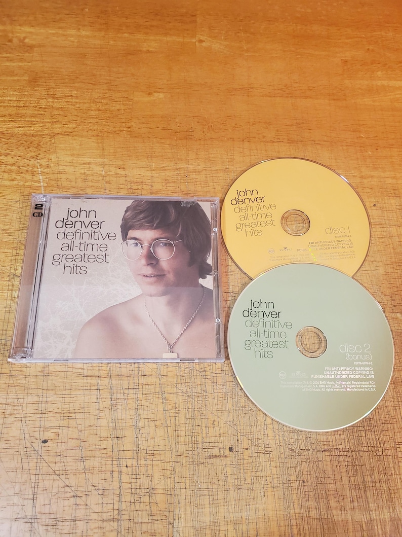 May include: A John Denver Definitive All-Time Greatest Hits 2-CD set in a clear plastic case. The cover features a portrait of John Denver wearing glasses and a necklace. Included are two discs, one yellow labelled disc 1 and one light green labelled disc 2.