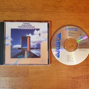 May include: A compact disc case and disc featuring "The Alan Parsons Project Instrumental Works." The CD case has a surreal landscape image with an open door. The disc is silver with blue text.