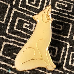 May include: A gold-colored pin in the shape of a howling wolf. The wolf is depicted in a stylized, minimalist design with a cream-colored enamel fill. The pin has a gold outline and is set against a black and white geometric patterned background.