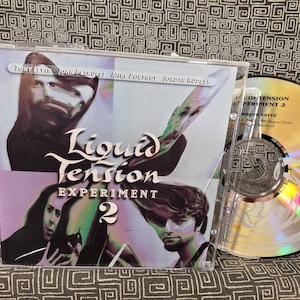 May include: A CD case with a purple and blue background featuring the band Liquid Tension Experiment 2. The band members are pictured on the front of the case. The CD is in a jewel case with a silver disc.