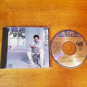 May include: A Lionel Richie Can't Slow Down compact disc and its plastic jewel case. The cover features a photograph of a person in a light blue shirt and white pants sitting on a chair. The disc displays the Motown logo and track list.