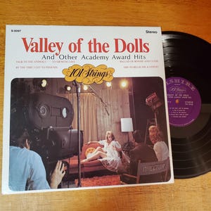 May include: A vintage vinyl record album titled "Valley of the Dolls" with "101 Strings" branding. The album cover features a film set scene with a woman in a white dress. A black vinyl record is visible next to the album cover.