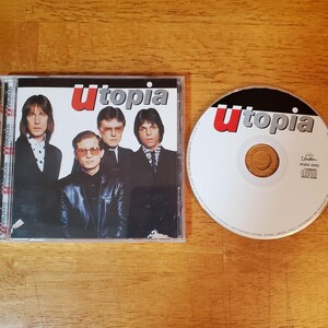 May include: A Utopia music CD in a clear plastic jewel case with a cover featuring four individuals in dark clothing. The disc itself is white with the red and black Utopia logo, the text AGEK-2050, and the Compact Disc Digital Audio symbol printed on it.