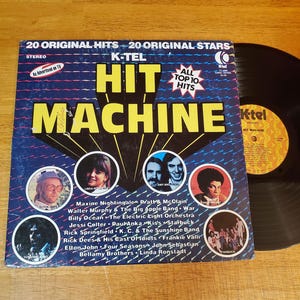 May include: A K-Tel Hit Machine vinyl record album cover featuring 20 original hits and stars. The blue cover displays yellow text, a starburst graphic, and circular portraits of artists including Elton John and Linda Ronstadt, with a black record.