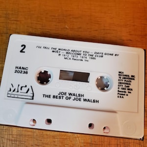K&ouml;nnte beinhalten: Eine wei&szlig;e Kassette mit dem Text "JOE WALSH THE BEST OF JOE WALSH" und dem MCA Records-Logo. Das Band enth&auml;lt Songtitel wie "I'LL TELL THE WORLD ABOUT YOU" und "WELCOME TO THE CLUB". Die Kassette ist mit "2" und "HANC 20236" beschriftet.