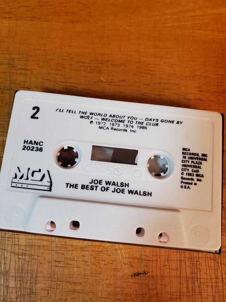 K&ouml;nnte beinhalten: Eine wei&szlig;e Kassette mit dem Text "JOE WALSH THE BEST OF JOE WALSH" und dem MCA Records-Logo. Das Band enth&auml;lt Songtitel wie "I'LL TELL THE WORLD ABOUT YOU" und "WELCOME TO THE CLUB". Die Kassette ist mit "2" und "HANC 20236" beschriftet.