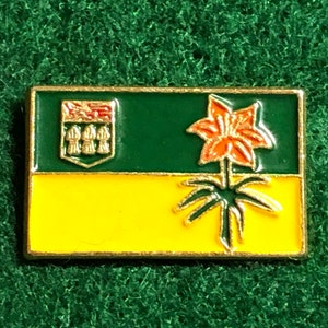 May include: A gold-colored pin with the flag of Saskatchewan. The flag features a green field with a yellow field below it. The green field has a crest with three gold wheat stalks and a red lily. The yellow field has a stylized orange lily.