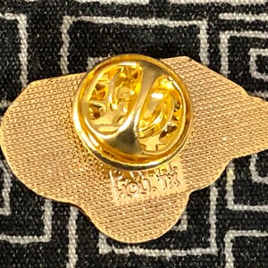 Puede incluir: Pin de metal dorado con una superficie texturizada y un cierre de pin. El pin tiene forma de cabeza de oso y tiene el texto "HOU. TX" en la parte posterior.