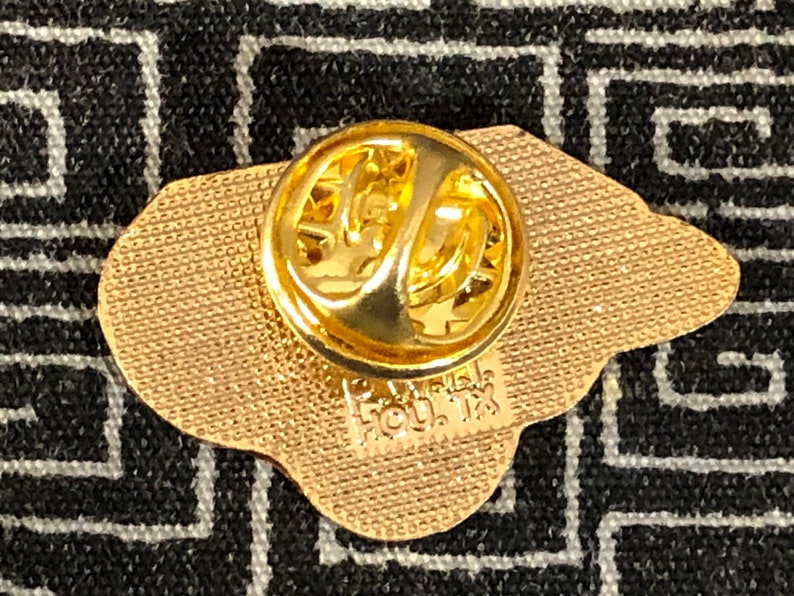 Puede incluir: Pin de metal dorado con una superficie texturizada y un cierre de pin. El pin tiene forma de cabeza de oso y tiene el texto "HOU. TX" en la parte posterior.