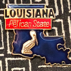 May include: A gold-colored pin shaped like the state of Louisiana. The pin has a blue background with a gold pelican on the left side. The top of the pin has a red banner with the words "Pelican State" in white letters. Above the banner is a black banner with the word "LOUISIANA" in white letters.