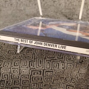 May include: A black CD case with a clear plastic cover. The front of the case has a photo of John Denver and the text "THE BEST OF JOHN DENVER LIVE" in white letters. The bottom right corner of the case has the word "LEGACY" in white letters.