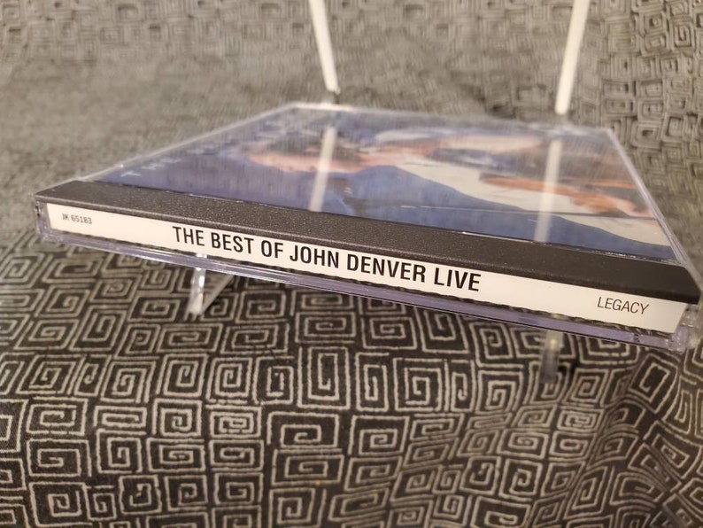 May include: A black CD case with a clear plastic cover. The front of the case has a photo of John Denver and the text "THE BEST OF JOHN DENVER LIVE" in white letters. The bottom right corner of the case has the word "LEGACY" in white letters.