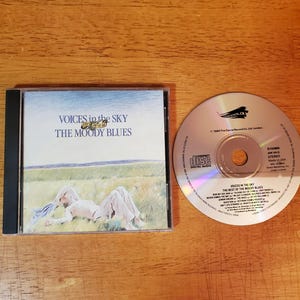 May include: A compact disc and its plastic jewel case for The Moody Blues album titled Voices in the Sky: The Best of The Moody Blues. The cover art features a person lying in a grassy field under a blue sky. The disc lists track titles.