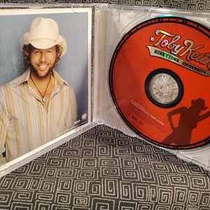 Może przedstawiać: Pudełko na płytę CD z czerwoną płytą CD z albumem Toby'ego Keitha Honky Tonk University. Okładka przedstawia sylwetkę kobiety w kowbojskim kapeluszu trzymającej pompon. Tylna okładka przedstawia zdjęcie Toby'ego Keitha w kowbojskim kapeluszu.