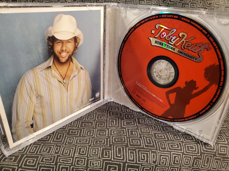 Może przedstawiać: Pudełko na płytę CD z czerwoną płytą CD z albumem Toby'ego Keitha Honky Tonk University. Okładka przedstawia sylwetkę kobiety w kowbojskim kapeluszu trzymającej pompon. Tylna okładka przedstawia zdjęcie Toby'ego Keitha w kowbojskim kapeluszu.