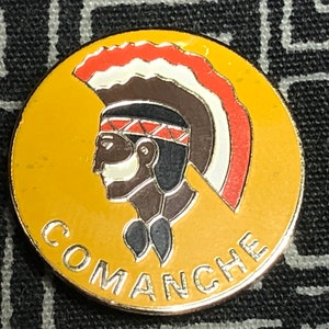 May include: A yellow metal pin with a stylized image of a Native American man wearing a headdress. The headdress is red, white, and black. The man's face is brown and he has a white stripe painted across his face. The pin says "COMANCHE" in silver lettering.