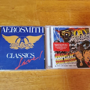 May include: Two compact discs, one blue with the text "Aerosmith Classics Live!" and the other with a colorful illustration of the band and the text "Music From Another Dimension!"
