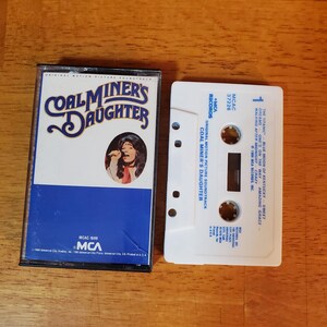 May include: A Coal Miner's Daughter original motion picture soundtrack cassette tape and its white plastic case. The case features blue and white text with a circular photograph of a singer. The white cassette tape lists track titles in blue.